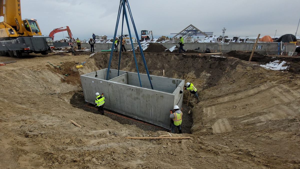 Crane lowering a large two-compartment precast grease interceptor into an excavation with crew guiding