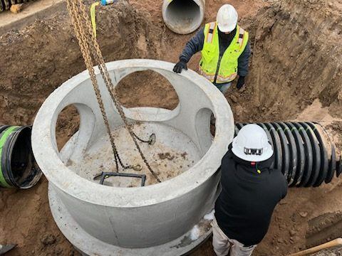 Crew lowering a precast concrete storm manhole barrel into the excavation